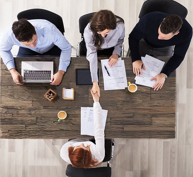 picture from above of four colleagues sitting at desk while two of them shake hands