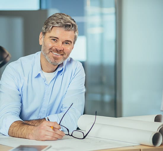 picture of man holding eye-glasses and sitting at desk with blueprints on it